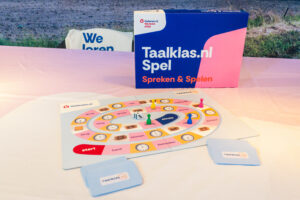 Colorful language game board on a white table with a blue-pink Taalklas.nl Spel box nearby and scattered cards.