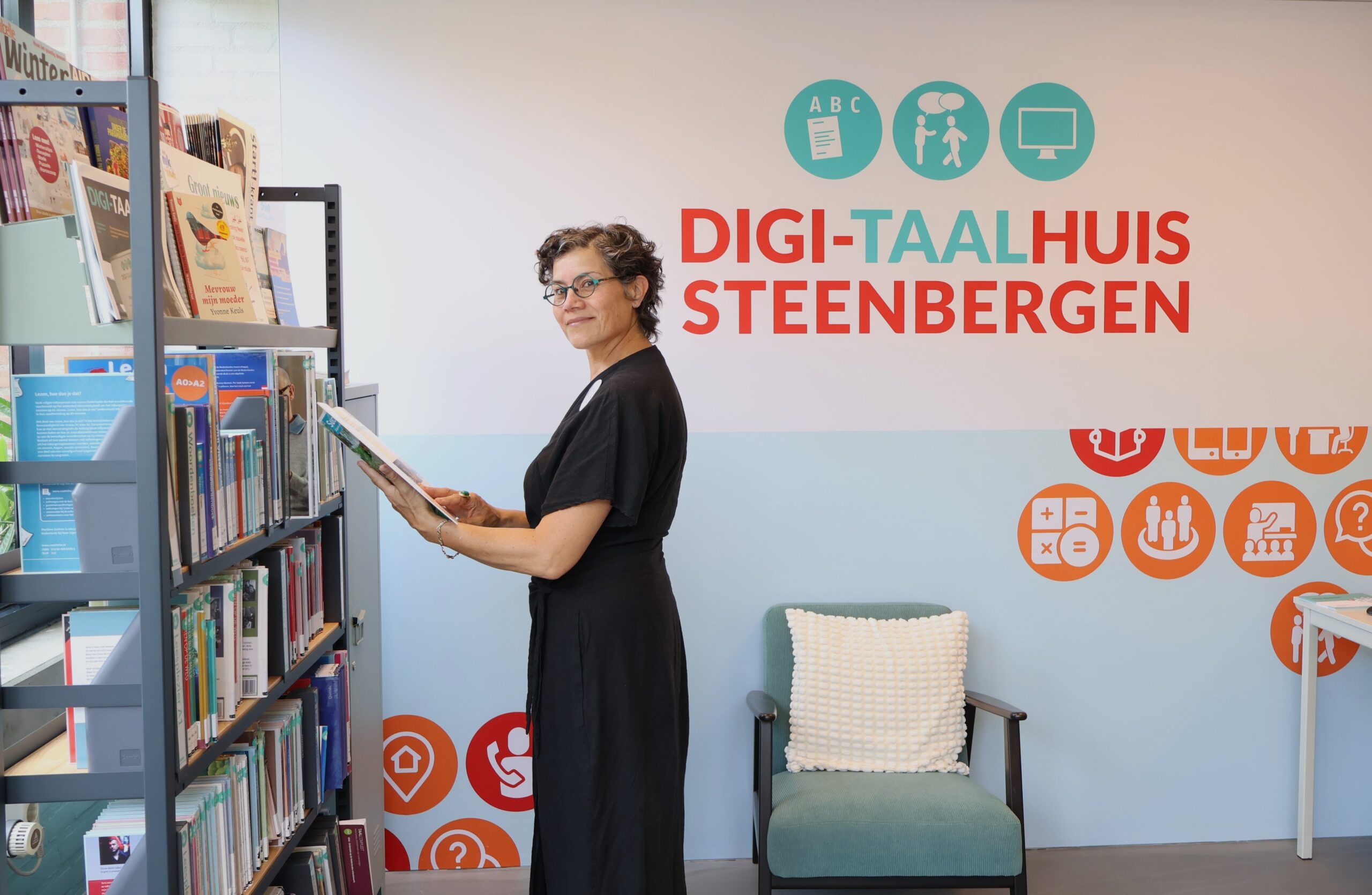 Woman in a black dress selecting a book from a library shelf, standing in front of a wall with Digi-Taalhuis Steenbergen branding and teal icons.