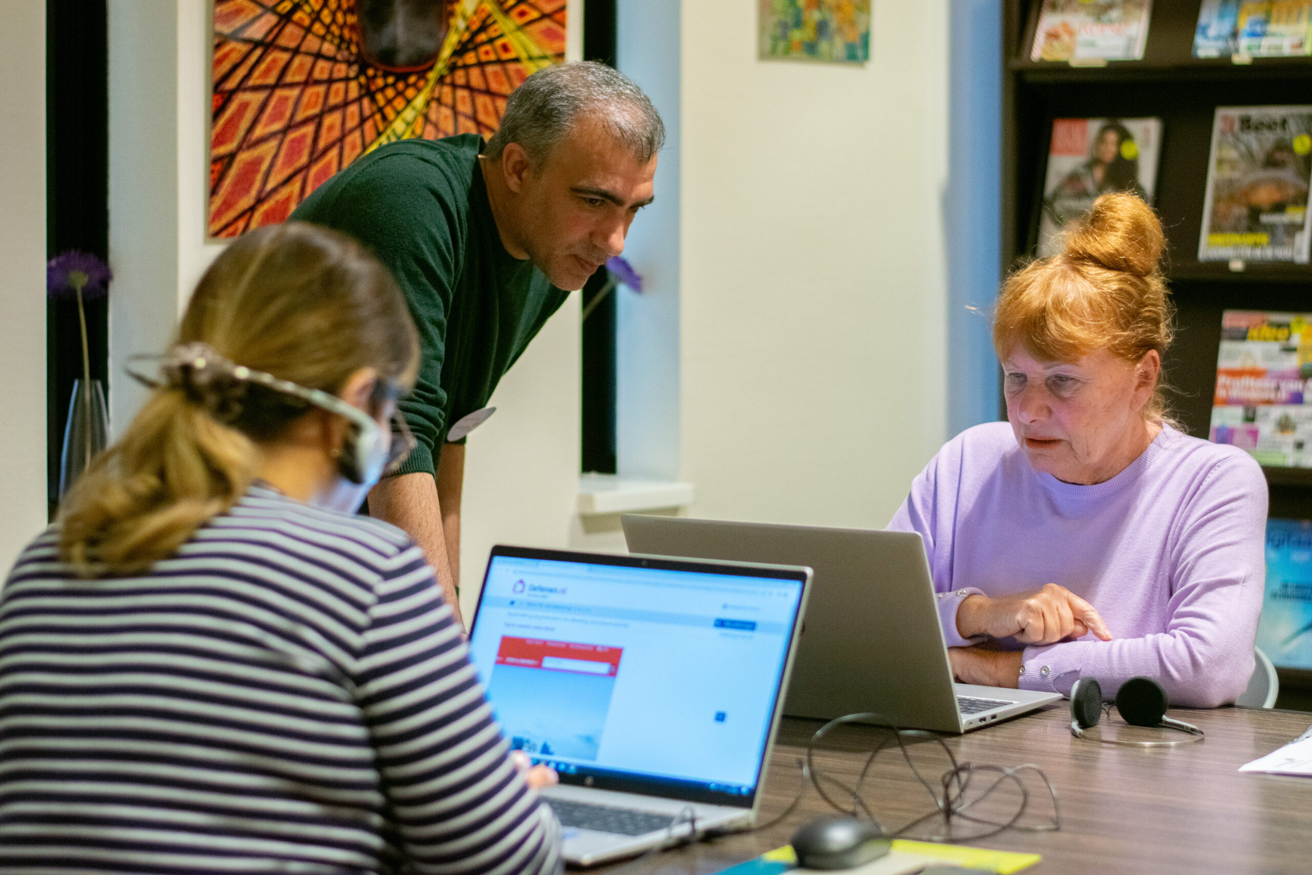 Group of adults collaborating around laptops at a table, with a man leaning over to help someone off-screen in a bright room with magazines on a shelf behind.
