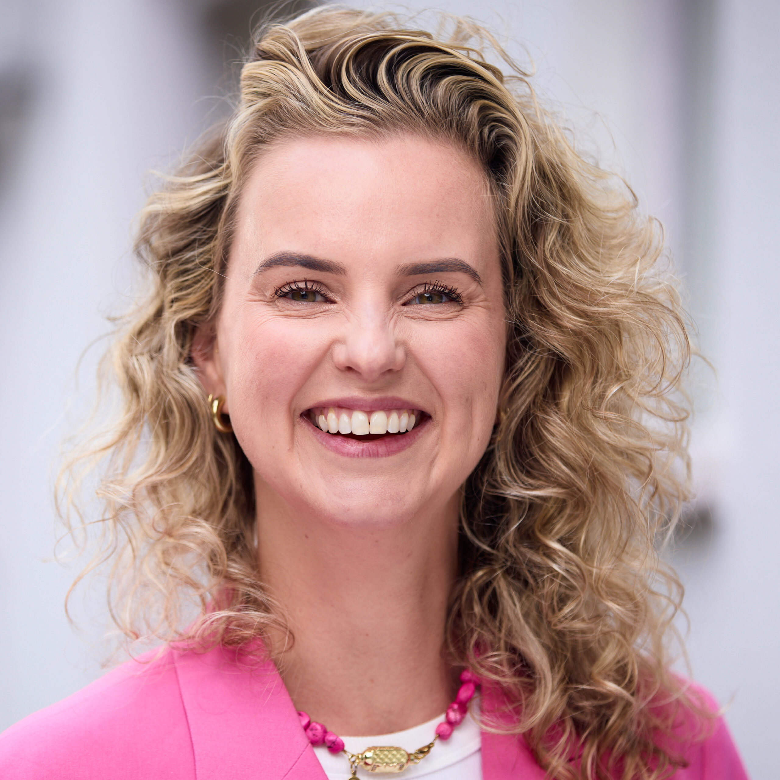 Close-up of a smiling woman with curly blonde hair wearing a bright pink blazer and pink beaded necklace.