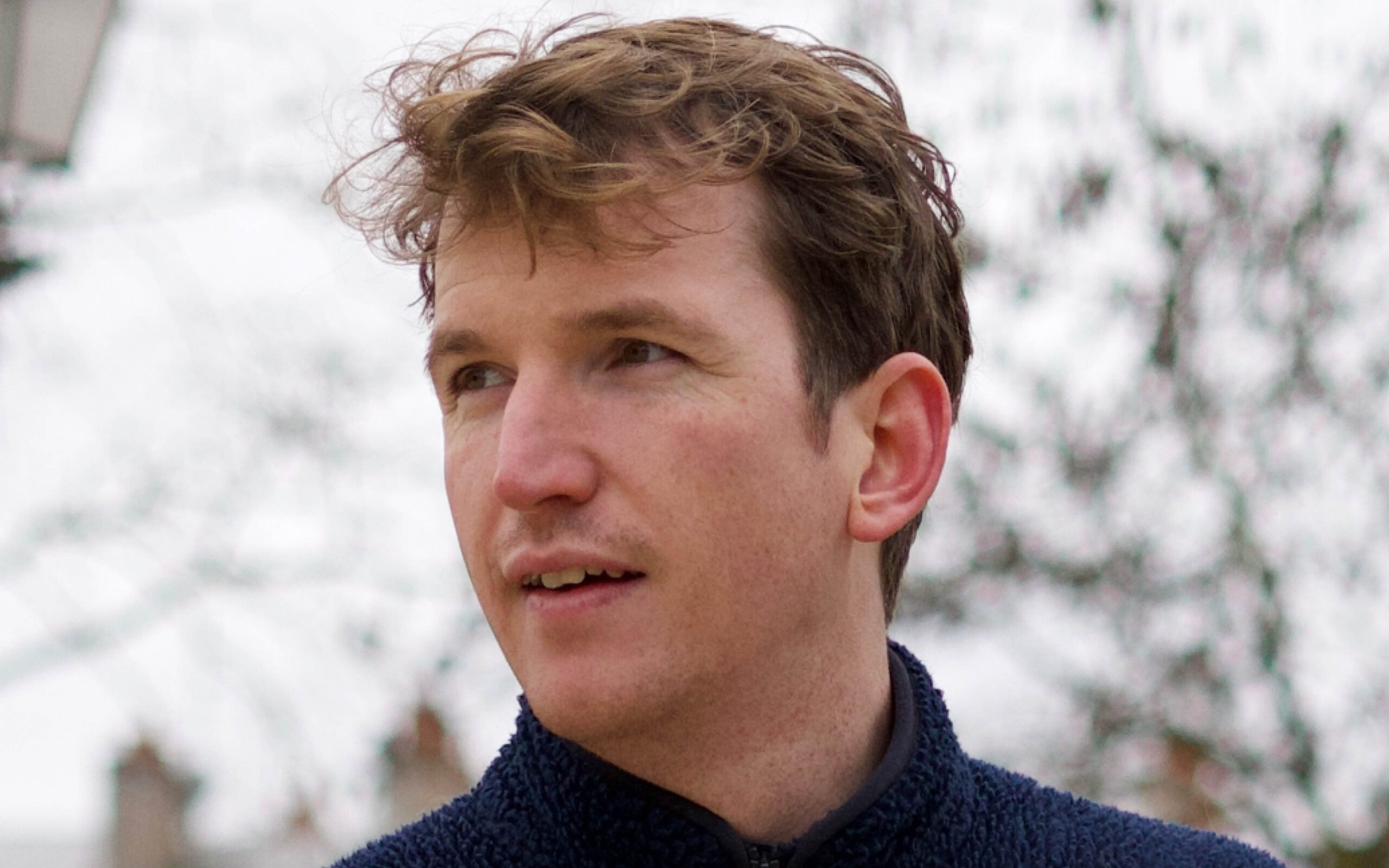 Close-up portrait of a young man outdoors, looking to the left, wearing a dark navy fleece.