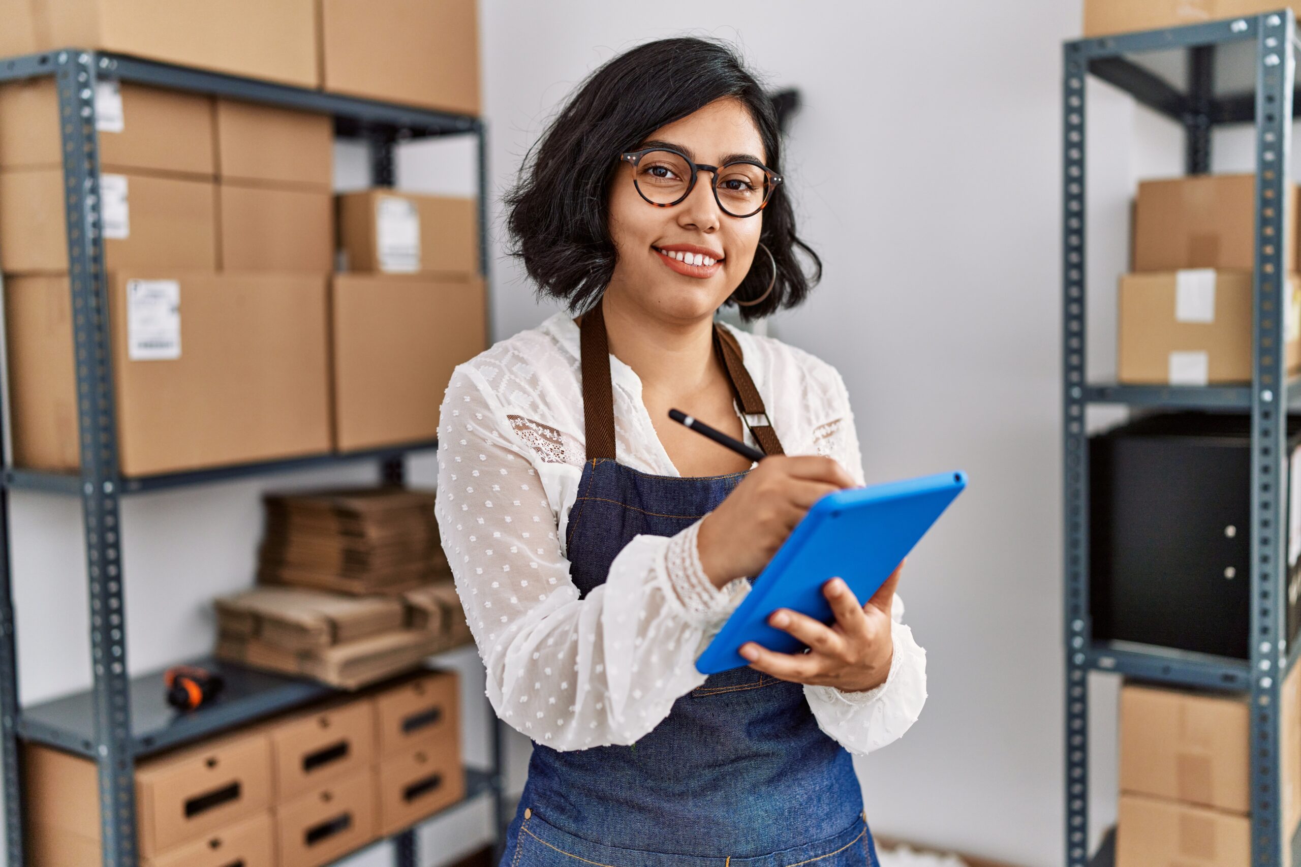 Smiling woman in a denim apron holding a blue clipboard in a warehouse with cardboard boxes on metal shelves behind her.