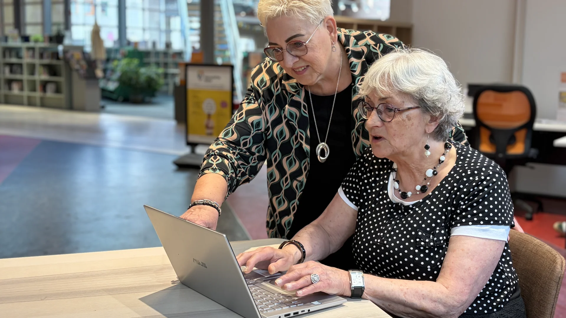 Two elderly women, one standing and guiding, look at a laptop together at a table in a bright library or cafe.